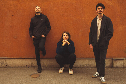 Jonatan Sirner trio poserar mot en orange vägg, en står lutad, en sitter på huk och en står rakt upp.