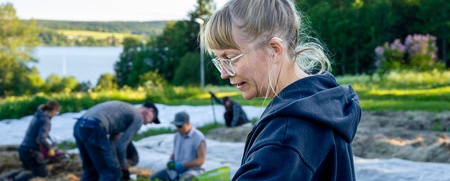 En kvinna med glasögon och uppsatt hår arbetar på en odlingsmark med andra personer i bakgrunden.