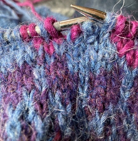 Närbild av stickning med blått och lila garn och stickor i metall.