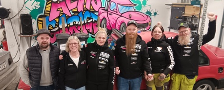 Sex personer står framför en röd bil i ett garage med färggraffiti på väggen.