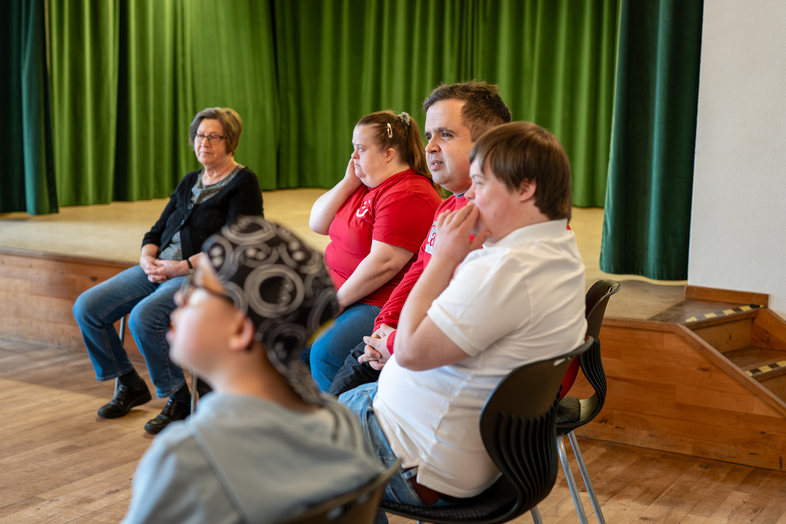 Fem personer sitter på stolar i en rad i ett rum med trägolv och grön scenbakgrund.