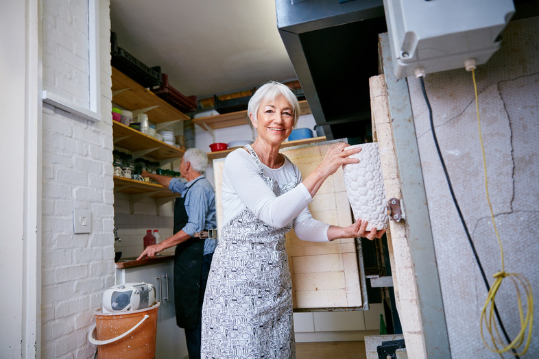 Äldre kvinna öppnar en keramikugn i en ljus verkstad, en man arbetar i bakgrunden.