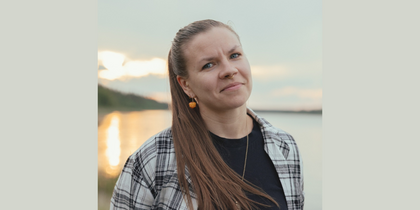 Person med långt hår och rutskjorta står utomhus vid en sjö i solnedgången.
