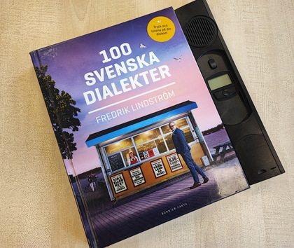Boken '100 svenska dialekter' av Fredrik Lindström ligger på ett bord bredvid en gammal bandspelare.