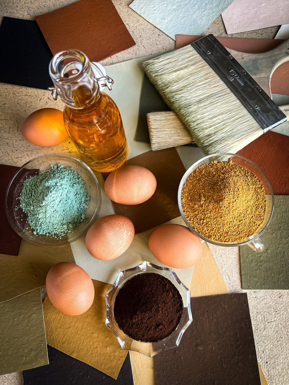 Flera ägg, glasburk med olja, färgpigment, penslar och färgprover på ett bord.