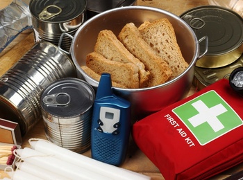 Nödutrustning med konserver, vattenflaskor, första hjälpen-kit, ficklampa, kniv, ljus och mediciner på ett träbord.
