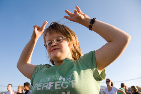 Ung person med glasögon och grön t-shirt dansar utomhus under blå himmel.