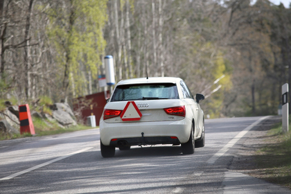 Vit Audi med övningskörningsskylt kör på landsväg omgiven av träd och vägmarkeringar.