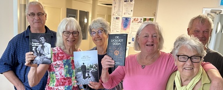 Sex äldre personer står tillsammans inomhus, tre av dem håller upp böcker och ler mot kameran.