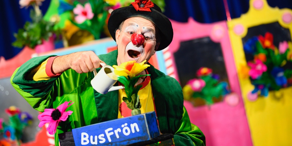 En person i färgglad clownkostym vattnar en blomma på en scen med färgglada dekorationer och skylten 'BusFrön'.