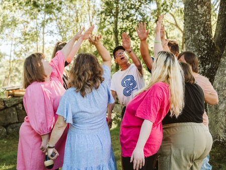 En grupp människor står i en ring utomhus och sträcker upp händerna tillsammans under träd.