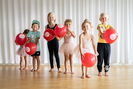 Sex glada barn står på rad inomhus med röda ballonger, några klädda i balettkläder.