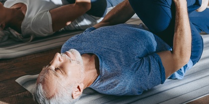 En grupp äldre personer gör liggande stretchövningar på yogamattor i ett ljust rum med trägolv.