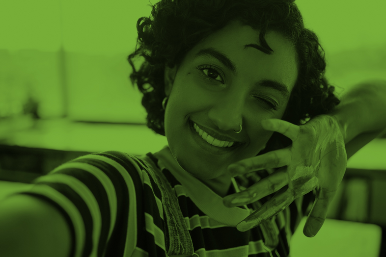 Person med lockigt hår tar en selfie, ler och visar en hand med färg på, i en inomhusmiljö med grönt filter.