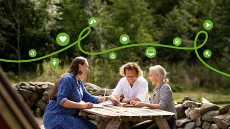 Tre personer sitter vid ett picknickbord utomhus och arbetar tillsammans med papper och dator.