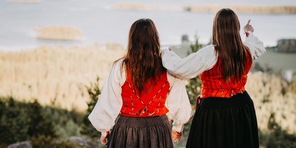Två personer i traditionella folkdräkter står på en klippa och ser ut över ett landskap med vatten och skog.