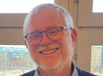 En person står inomhus framför stora fönster med utsikt över en stadsmiljö och parkerade bilar.