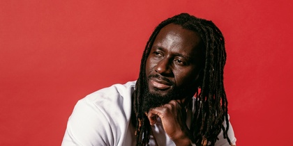Pa Modou Badjie i vit t-shirt och svarta arbetsbyxor sitter på högtalare mot röd bakgrund