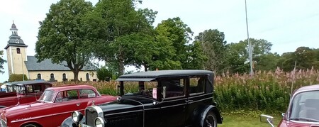 Flera klassiska bilar står parkerade på en gräsmatta med en kyrka och träd i bakgrunden.