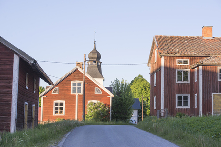Landsväg mellan röda trähus och en kyrkobyggnad med torn i en lantlig miljö.