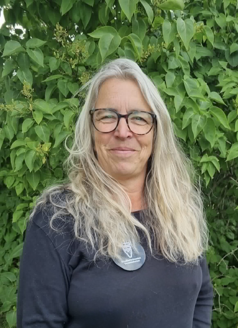Kvinna med långt blont hår och glasögon står framför grönskande buskar utomhus.