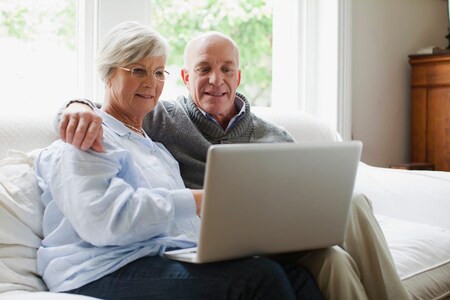 En man och en kvinna sitter i en vit soffa och tittar på en laptop. Mannen håller sin arm om kvinnans axlar.