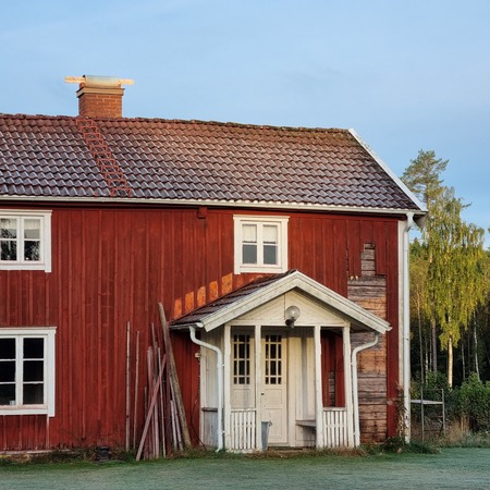 Röd trähus med vita detaljer och tegeltak, omgiven av grönska och morgondagg på marken.
