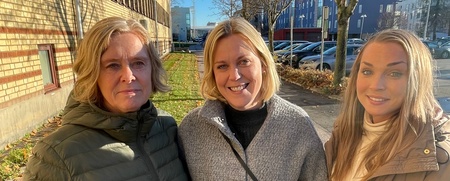 Tre kvinnor står tillsammans utomhus i solsken, klädda i varma jackor, med byggnader och träd i bakgrunden.