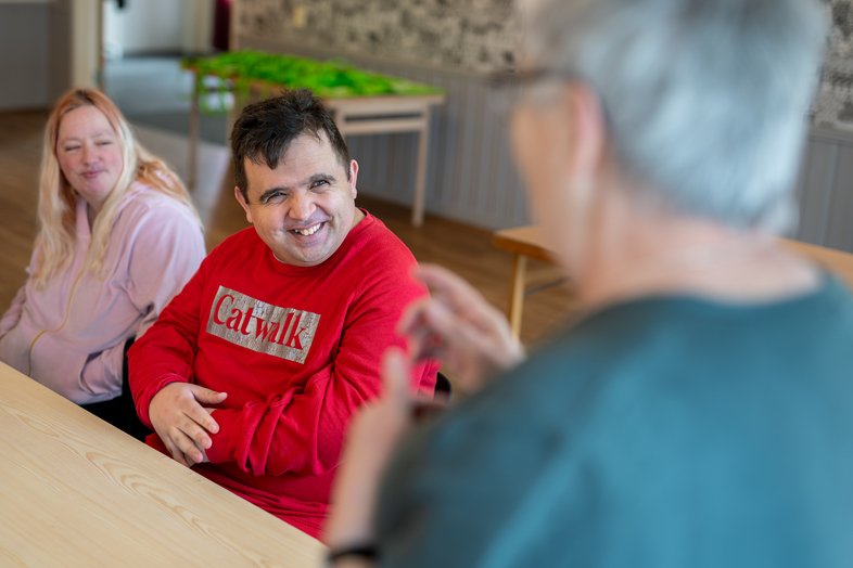 Två personer sitter vid ett bord och lyssnar på en tredje person som pratar framför dem.
