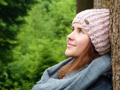 Person med stickad mössa och halsduk lutar mot ett träd i en grön skogsmiljö.