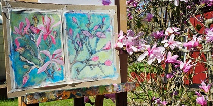 Ett staffli med målningar av rosa magnoliablommor står utomhus bredvid ett blommande magnoliaträd.