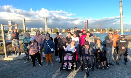 En grupp människor, inklusive personer i rullstol, poserar utomhus vid vattnet under en solig dag.