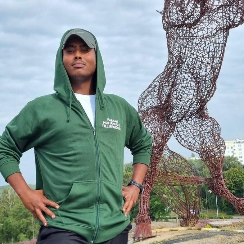 En person i grön huvtröja står utomhus framför en stor metallskulptur av en mänsklig figur.