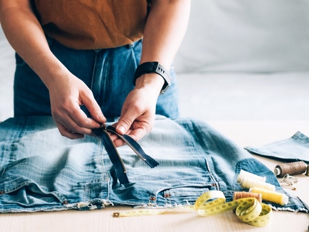 Person arbetar med att sy om ett par jeans, med dragkedja, måttband och sytråd på bordet.