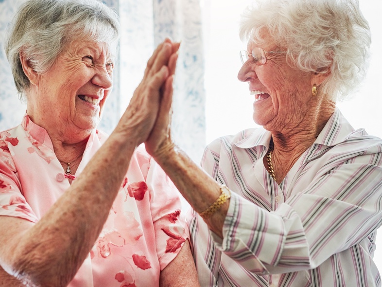 Två äldre kvinnor skrattar och ger varandra en high five i ett ljust rum.