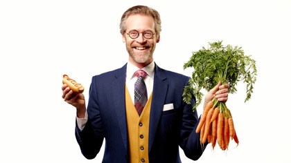 Man i kostym håller en varmkorv i ena handen och en bunt morötter i den andra.