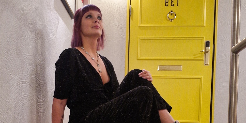 Person sitter avslappnat på en trappa framför en gul dörr, klädd i svart dräkt och svarta Converse-skor.