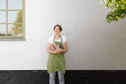 Person i grön förkläde håller bröd framför en vit vägg, ansiktet är suddat.