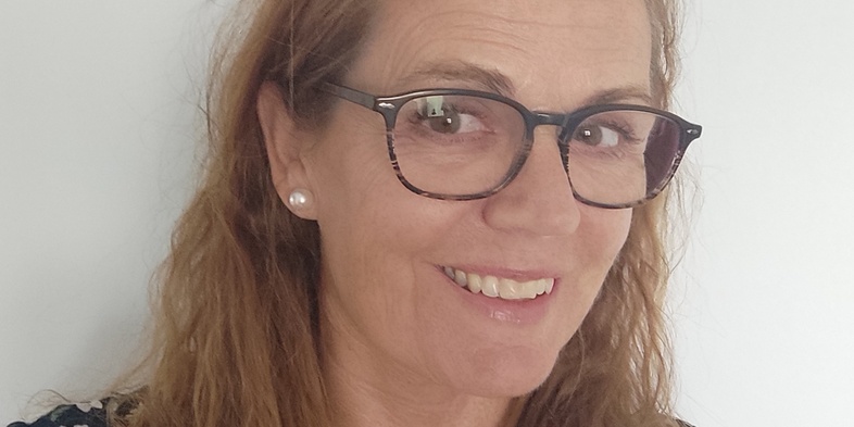 Kvinna med långt, vågigt hår och glasögon ler mot kameran framför en vit bakgrund.