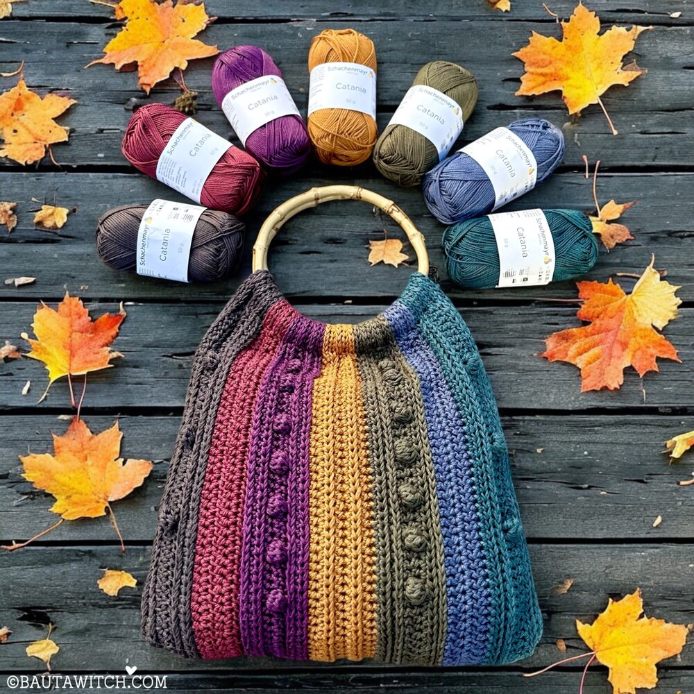 Virkad väska i regnbågsfärger med garnnystan och höstlöv på ett träbord.