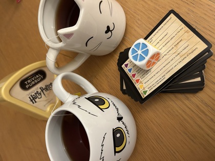 Två tekoppar med uggle- och kattmotiv, Harry Potter Trivial Pursuit, spelkort och tärning på ett träbord.
