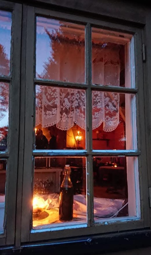 Vy genom ett fönster in i ett mysigt rum med spetsgardiner, tända ljus och en flaska på bordet.