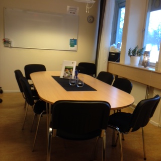 Ett litet mötesrum med ett ovalt bord, åtta svarta stolar, whiteboard och fönster med växter.