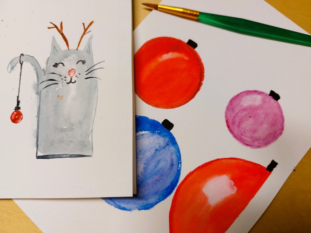 Två akvarellmålningar: en katt med renhorn och julkula, samt färgglada julgranskulor och en pensel.