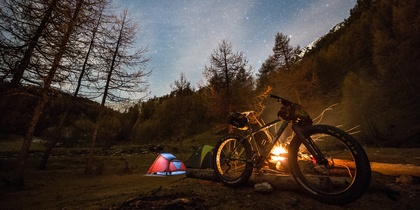 Två tält och en cykel vid en lägereld under en stjärnklar natthimmel i en skog.