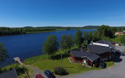 En röd byggnad vid en flod, omgiven av träd och bilar, under en klarblå himmel.