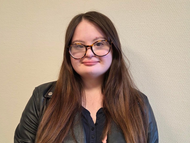 Person med långt brunt hår och svart skinnjacka står mot en ljus vägg. Ansiktet är suddat.