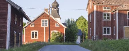 Landsväg mellan röda trähus och en kyrkobyggnad med torn i en lantlig miljö.