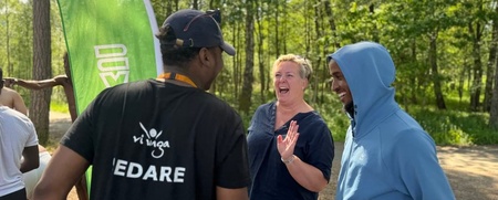 Tre personer samtalar och skrattar utomhus vid en grön flagga i en solig skogsmiljö.