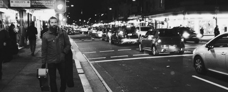 Svartvit nattbild av en stadsgata med bilar och en person som går på trottoaren.
